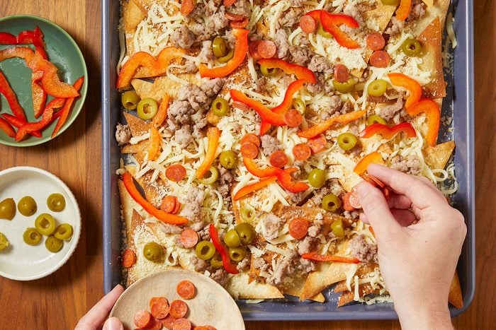 Overhead horizontal image of nachos being assembled in a baking pan. Sliced pepperoni, red bell peppers, olives, cooked ground meat and shredded cheese are being layered on top of the tortilla chips.