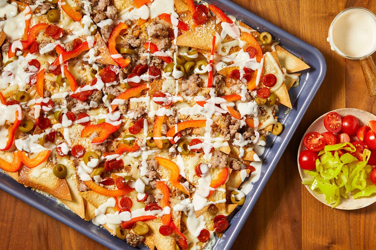 Overhead horizontal image of loaded nachos on a sheet pan with a light sauce drizzled on top. Nachos include sausage, olives, pepperoni, and sliced red peppers, with a side of tomatoes and banana peppers.