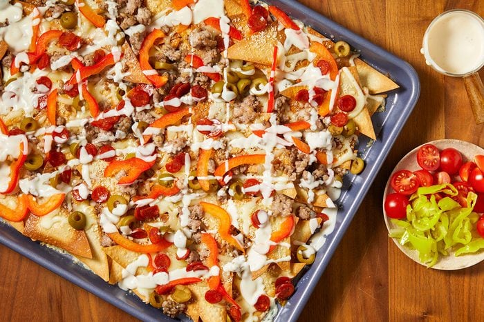 Overhead horizontal image of loaded nachos on a sheet pan with a light sauce drizzled on top. Nachos include sausage, olives, pepperoni, and sliced red peppers, with a side of tomatoes and banana peppers.
