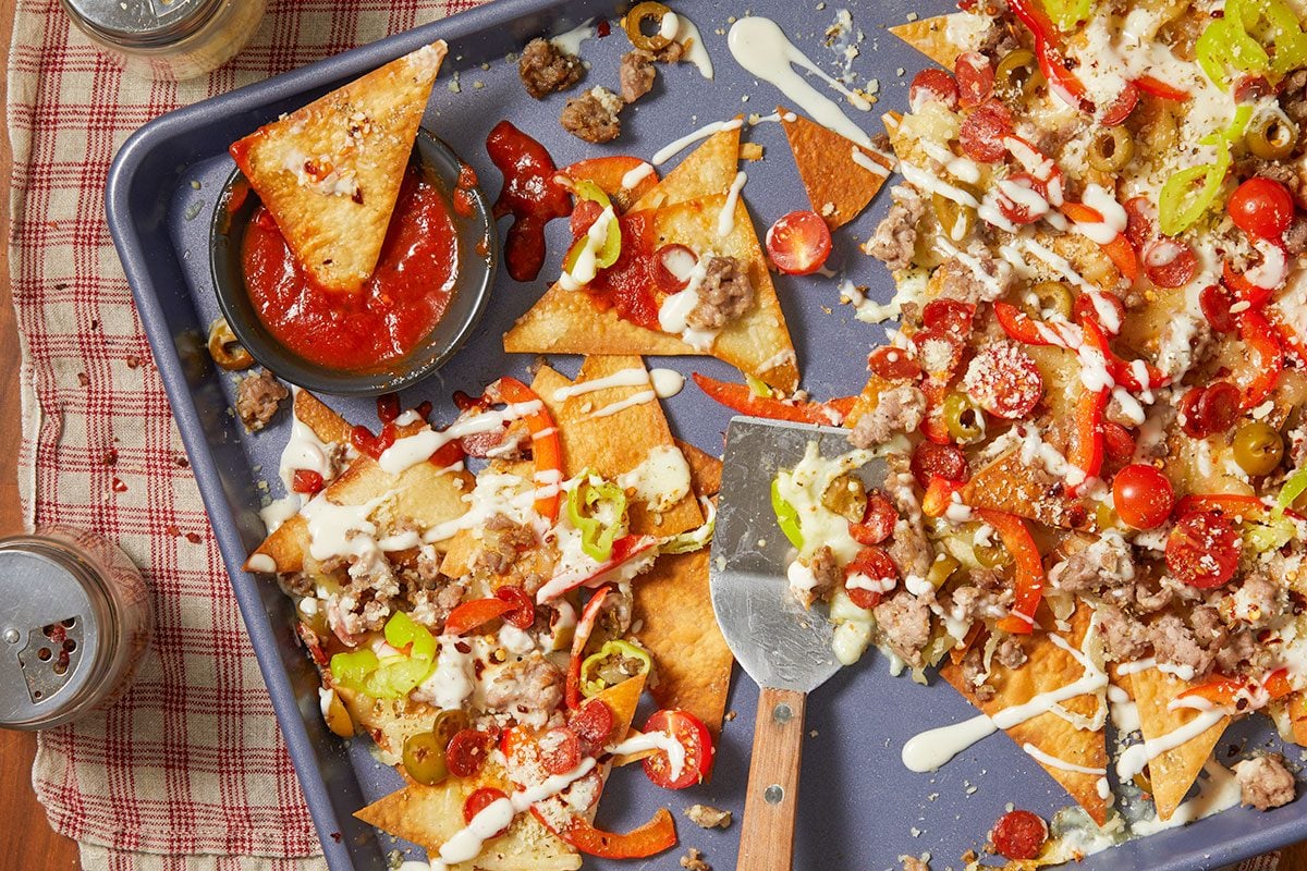 Overhead horizontal image of pizza nachos on a blue baking sheet. The image shows the pizza nachos being assembled, topped with ground sausage, pepperoni, tomatoes, olives, green peppers, and a creamy sauce. A bowl of marinara sauce sits on the side.