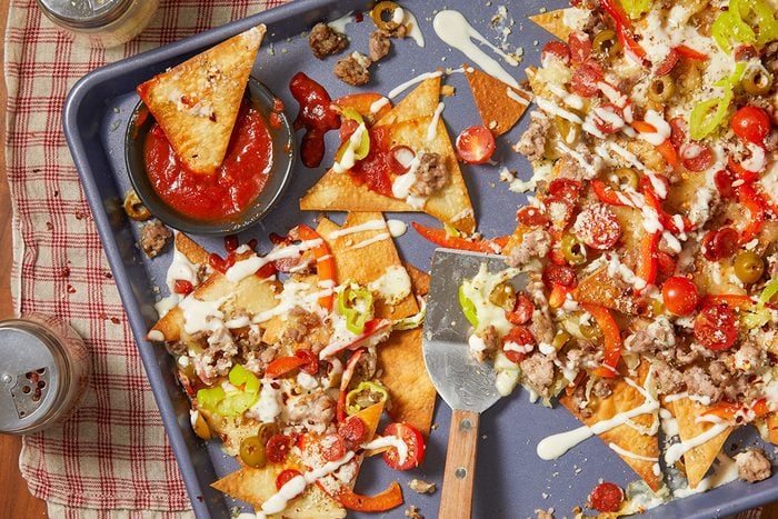 Overhead horizontal image of pizza nachos on a blue baking sheet. The image shows the pizza nachos being assembled, topped with ground sausage, pepperoni, tomatoes, olives, green peppers, and a creamy sauce. A bowl of marinara sauce sits on the side.