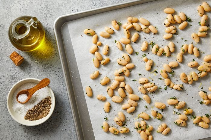 Overhead horizontal image of white beans scattered on a parchment paper lined baking sheet. A bowl of salt and pepper with a small wooden spoon and a bottle of olive oil sit next to the tray, on a grey background.