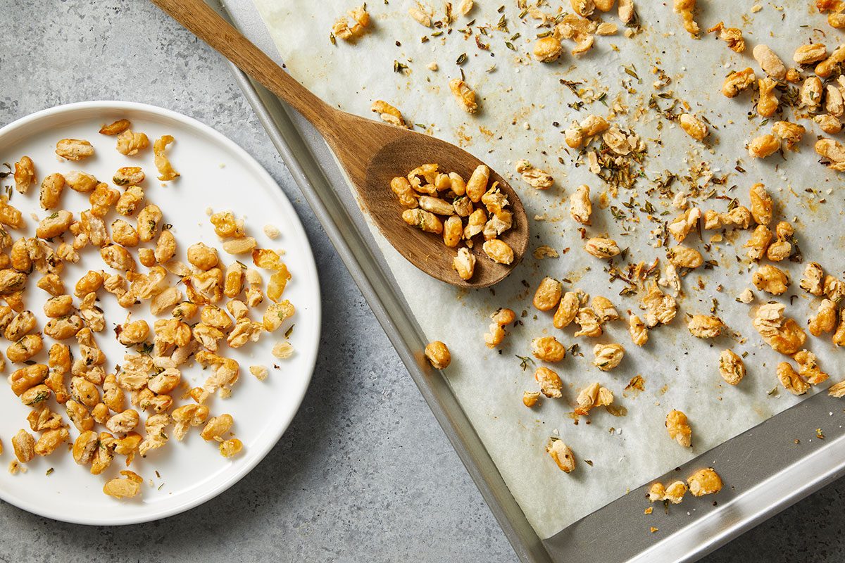 Overhead horizontal image of seasoned peanuts being transferred from a parchment-lined baking sheet using a wooden spoon onto a white plate. The baking sheet has peanuts and some herbs scattered across it.
