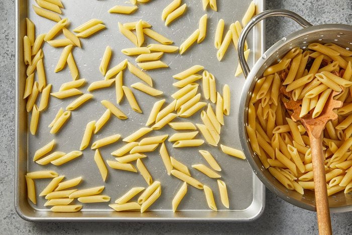 Overhead horizontal image of penne pasta. Part of the cooked pasta sits inside a colander with a wooden spoon, and part of the pasta sits spread out on a baking sheet.
