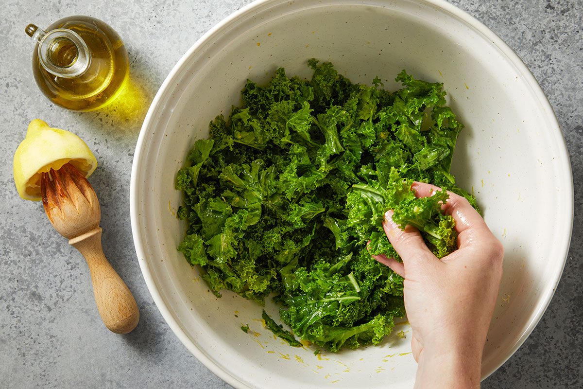 Overhead horizontal image of a white bowl filled with chopped kale being held in a person's hand. A bottle of olive oil and lemon squeezer with lemon are on the side.