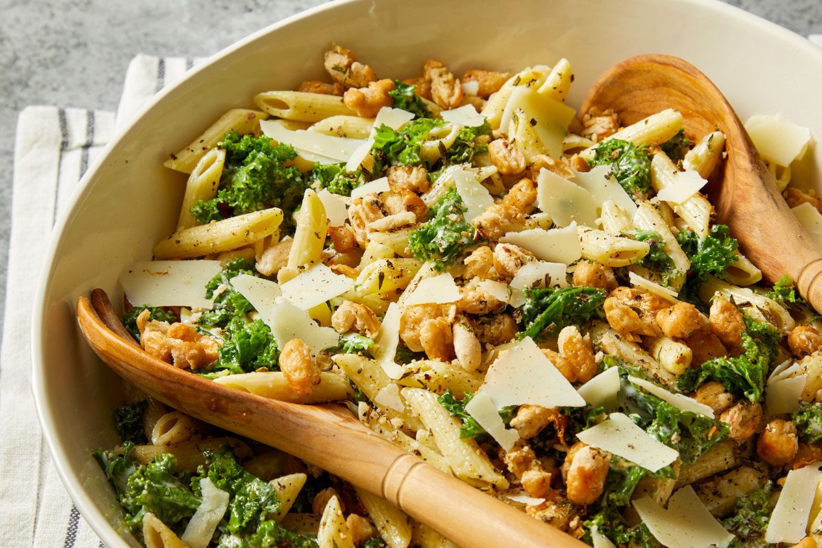 Overhead horizontal image of pasta salad with kale, wooden salad spoons, crispy chickpeas and Parmesan cheese in a white bowl. The salad is on a striped napkin and gray background.