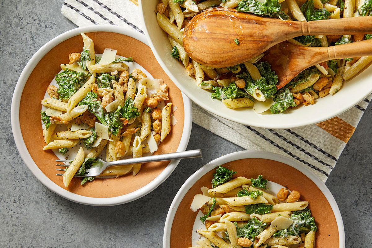 Overhead horizontal image of pasta salad in three dishes. The dish in the upper right is a large serving bowl of the finished pasta salad. The salad includes pasta, kale, chickpeas and parmesan cheese.
