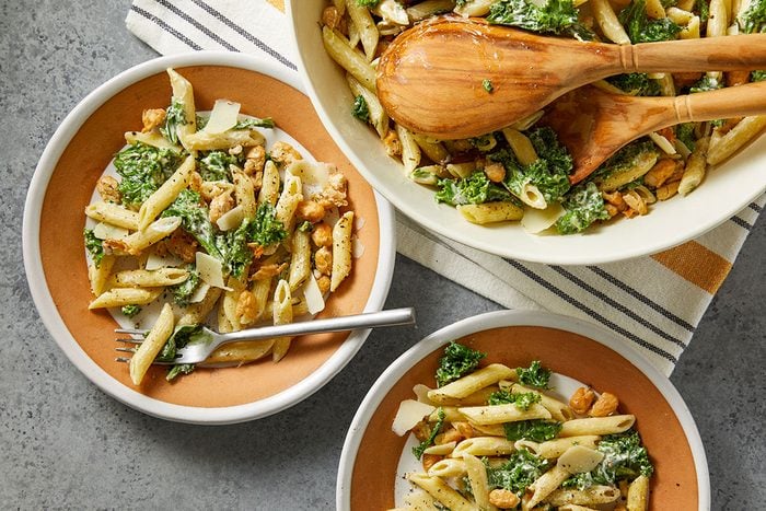 Overhead horizontal image of pasta salad in three dishes. The dish in the upper right is a large serving bowl of the finished pasta salad. The salad includes pasta, kale, chickpeas and parmesan cheese.