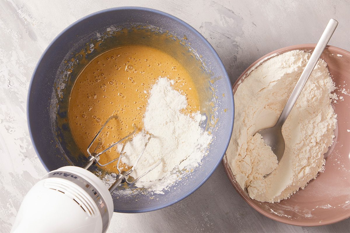 Overhead horizontal image of batter in a blue bowl with white flour being added using a hand mixer. Next to the bowl is a pink plate with a mound of flour and a metal spoon.