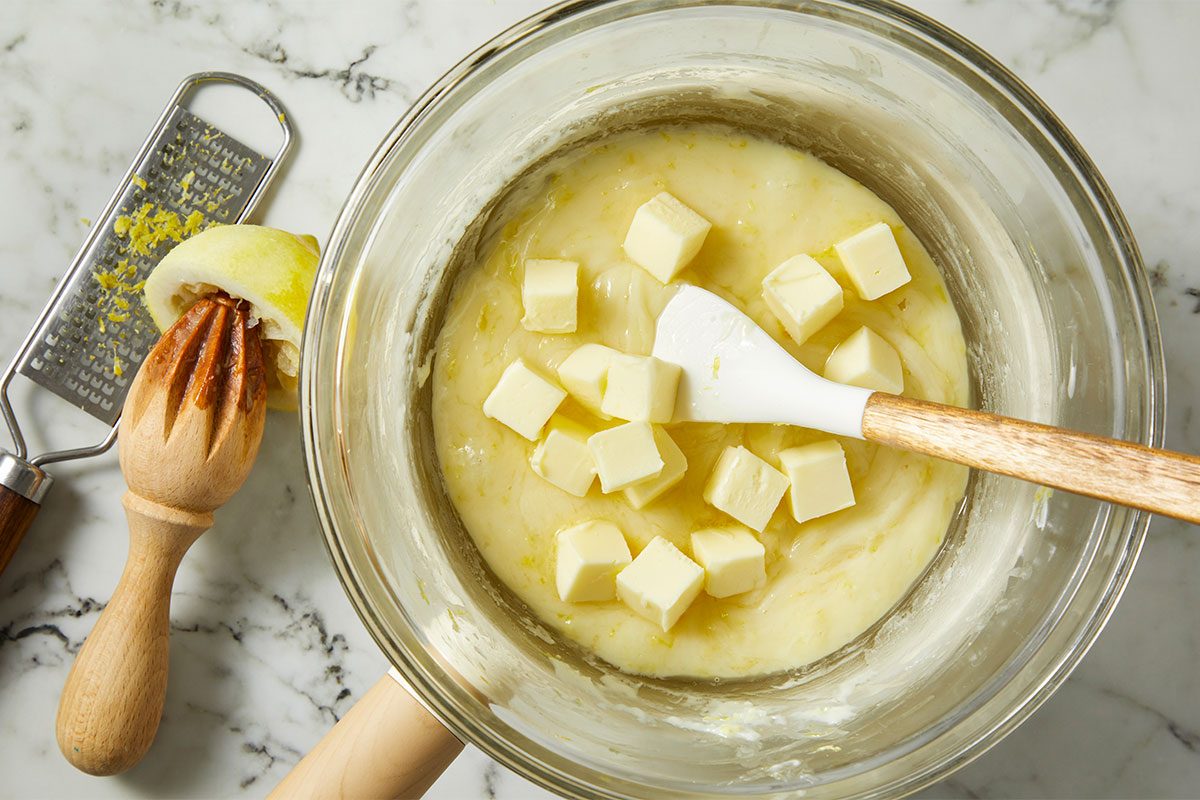 A glass bowl with a creamy mixture and cubes of butter, stirred with a spatula. Nearby are a citrus reamer with a squeezed lemon half and a grater with lemon zest on a marble countertop.