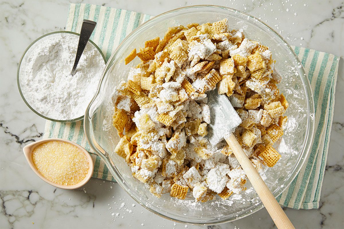 A glass bowl of powdered sugar-coated Chex cereal being mixed with a spatula, next to bowls of powdered sugar and yellow sprinkles on a green-striped cloth atop a marble surface.