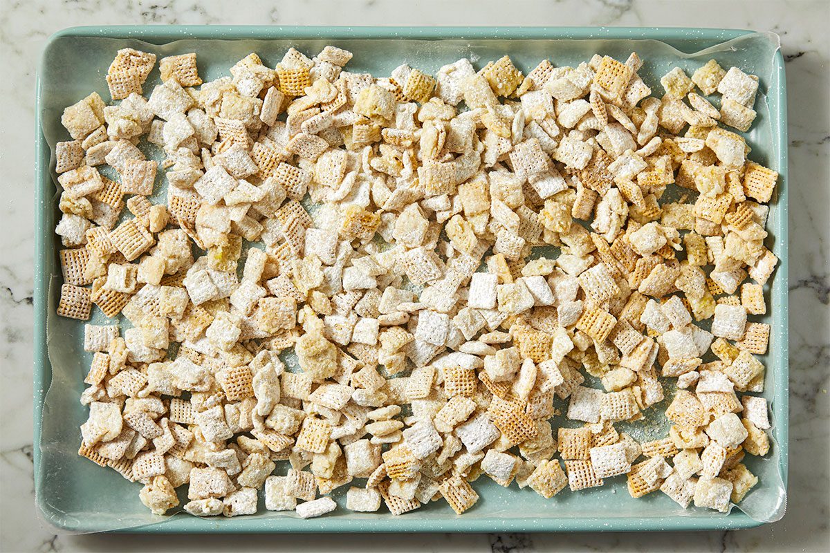 A baking sheet is covered with a single layer of Chex cereal pieces coated in powdered sugar, likely prepared for a snack such as Puppy Chow or Muddy Buddies. The tray rests on a marble surface.