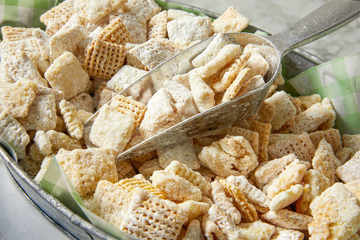 A metal scoop rests in a tray filled with powdered sugar-coated cereal squares, likely homemade puppy chow or Muddy Buddies, on a green and white checkered paper liner.