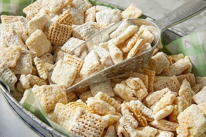 A metal scoop rests in a tray filled with powdered sugar-coated cereal squares, likely homemade puppy chow or Muddy Buddies, on a green and white checkered paper liner.