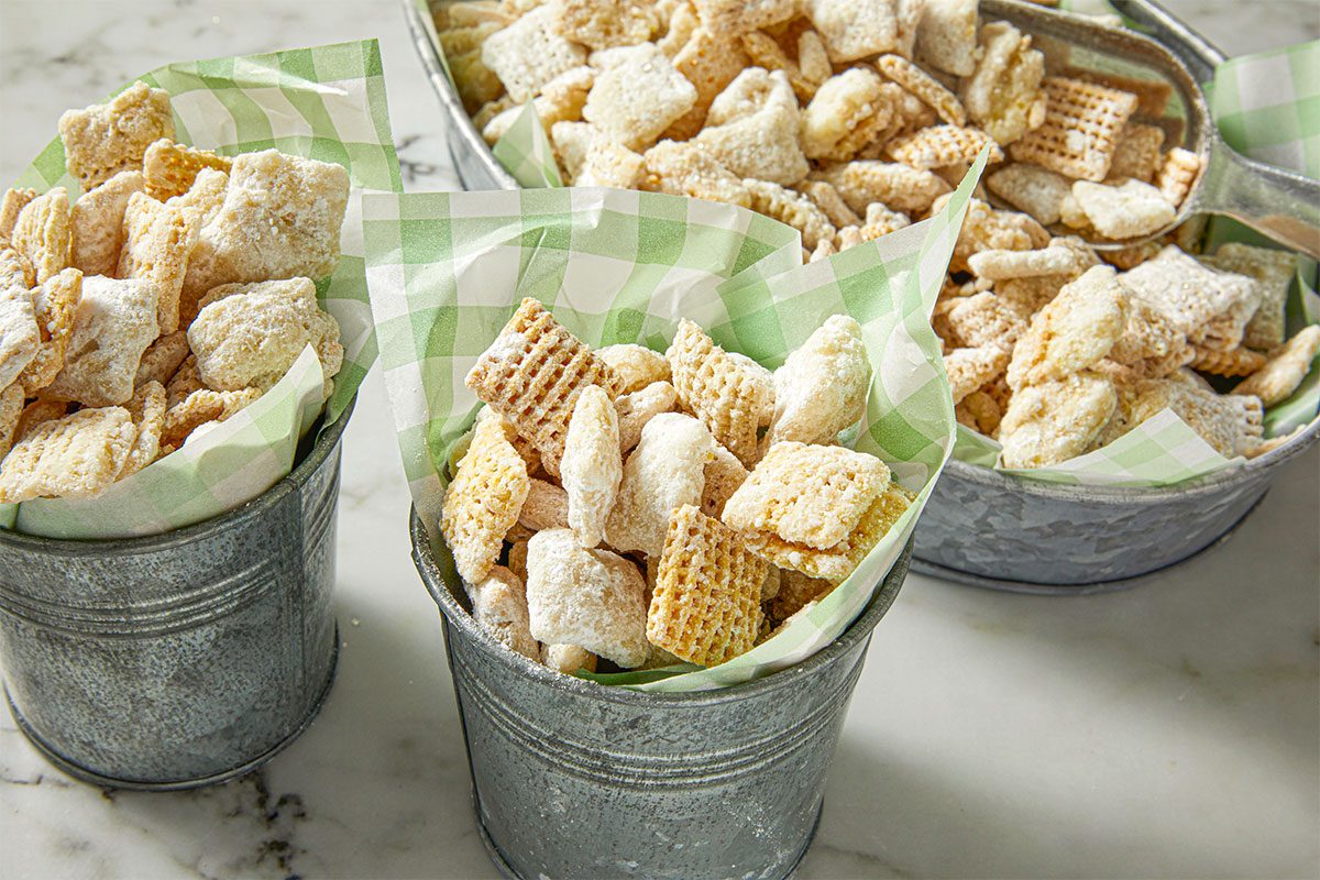 Small metal buckets lined with green checkered paper, each filled with Chex mix coated in powdered sugar, sit on a marble surface. The snack mix features square cereal pieces and is served in a casual, rustic style.