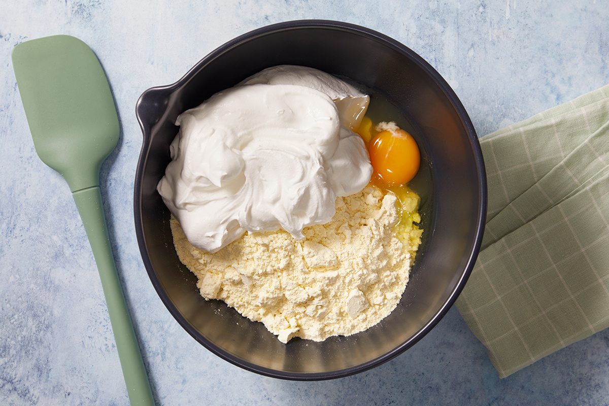 A black mixing bowl containing flour, a mound of whipped egg whites, a whole egg, and some liquid, sits on a light countertop next to a green spatula and a folded green kitchen towel.