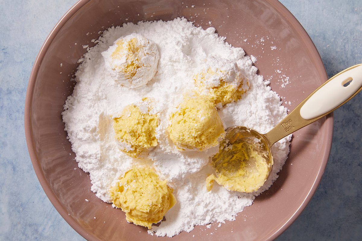 A mixing bowl filled with powdered sugar and several scoops of yellow cookie dough, with a spoon resting inside the bowl.