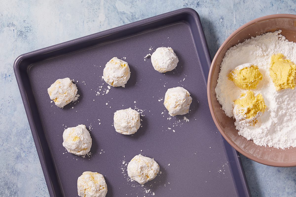 A baking tray with eight unbaked dough balls coated in powdered sugar sits next to a bowl containing dough and more powdered sugar on a light blue surface.