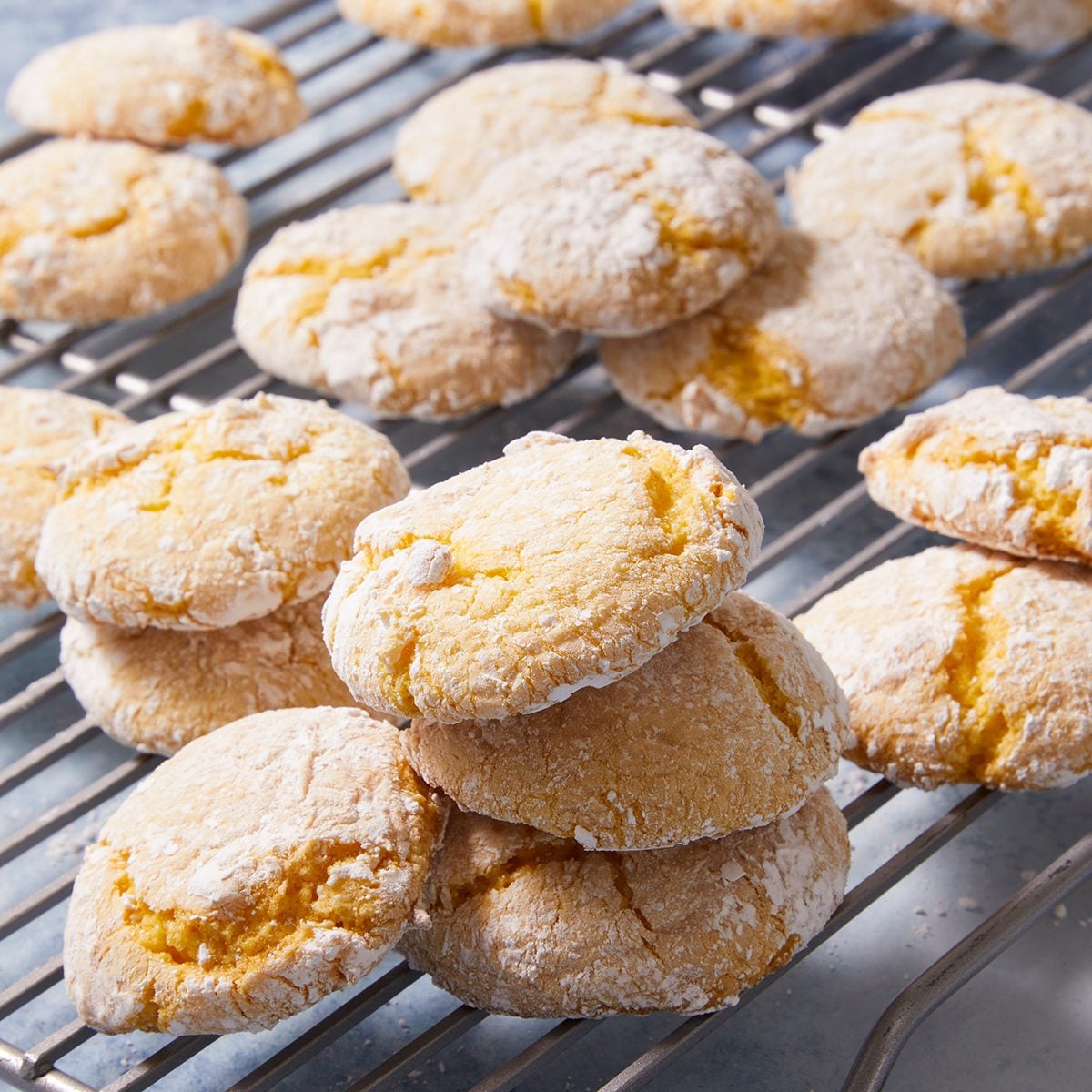 A stack of golden, cracked amaretti cookies dusted with powdered sugar is displayed on a cooling rack, with more cookies scattered in the background.