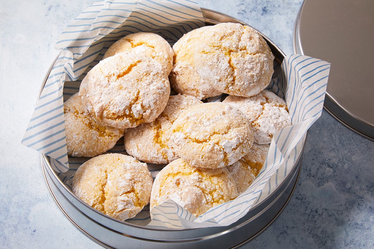 A round tin lined with striped parchment paper is filled with several powdered sugar-covered cookies, arranged in a casual pile. The tin sits on a light-colored surface.