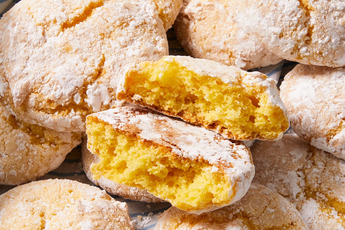 A close-up of several powdered sugar-covered cookies, with one cookie broken in half to show its soft, yellow, airy interior.
