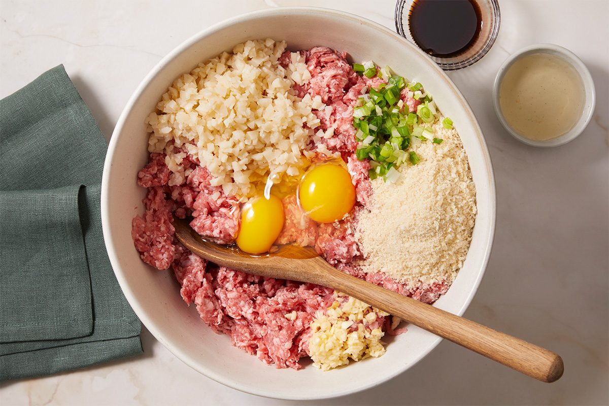 Overhead horizontal image of various ingredients in a white bowl, including ground meat, eggs, rice, scallions, and parmesan cheese. A wooden spoon rests inside the bowl of ingredients.