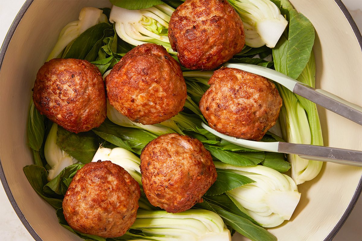 Overhead horizontal image of cooked meatballs arranged on a bed of bok choy with tongs. The meatballs are browned and positioned around fresh green bok choy.