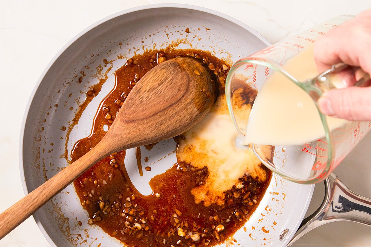 Overhead horizontal image of an off-white pan with sauteed garlic and a wooden spoon. A hand is pouring a light yellow liquid into the pan to create a sauce.