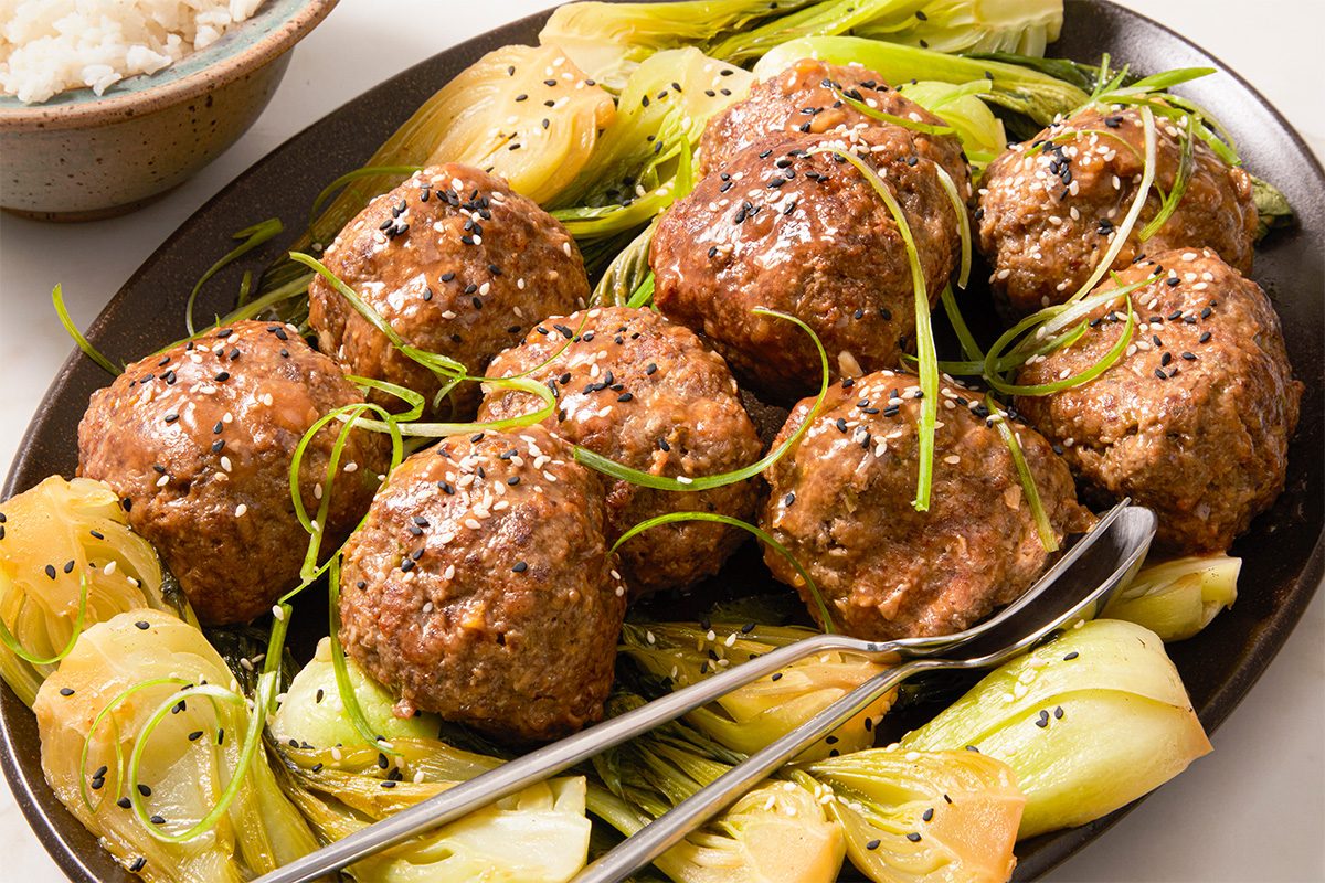 Eye Level horizontal image of a serving platter with multiple meatballs. The meatballs are topped with sesame seeds and green onions, and surrounded by bok choy. A serving of rice is in a bowl in the background.