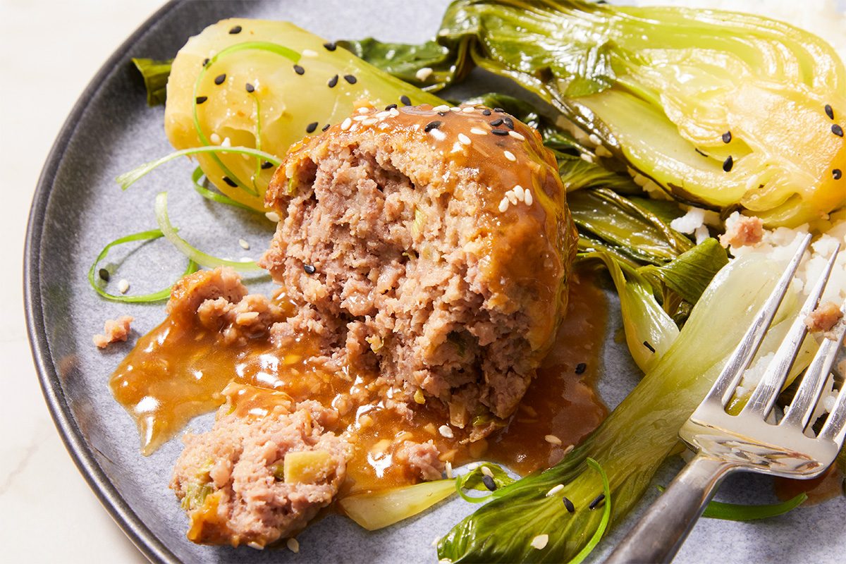 Eye Level horizontal image of a partially cut meatball on a grey plate, covered in a brown sauce and garnished with sesame seeds. Steamed bok choy is visible in the background, and there's a fork on the side.