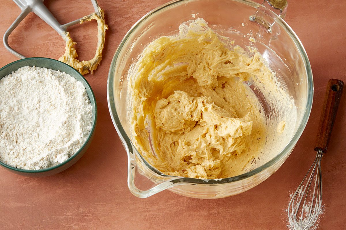 Overhead horizontal image of batter being mixed in a clear glass bowl, with a mixer attachment and a whisk laying nearby. There is a bowl of flour next to the mixer attachment. | [Original] Adding flour mixture to creamed butter mixture