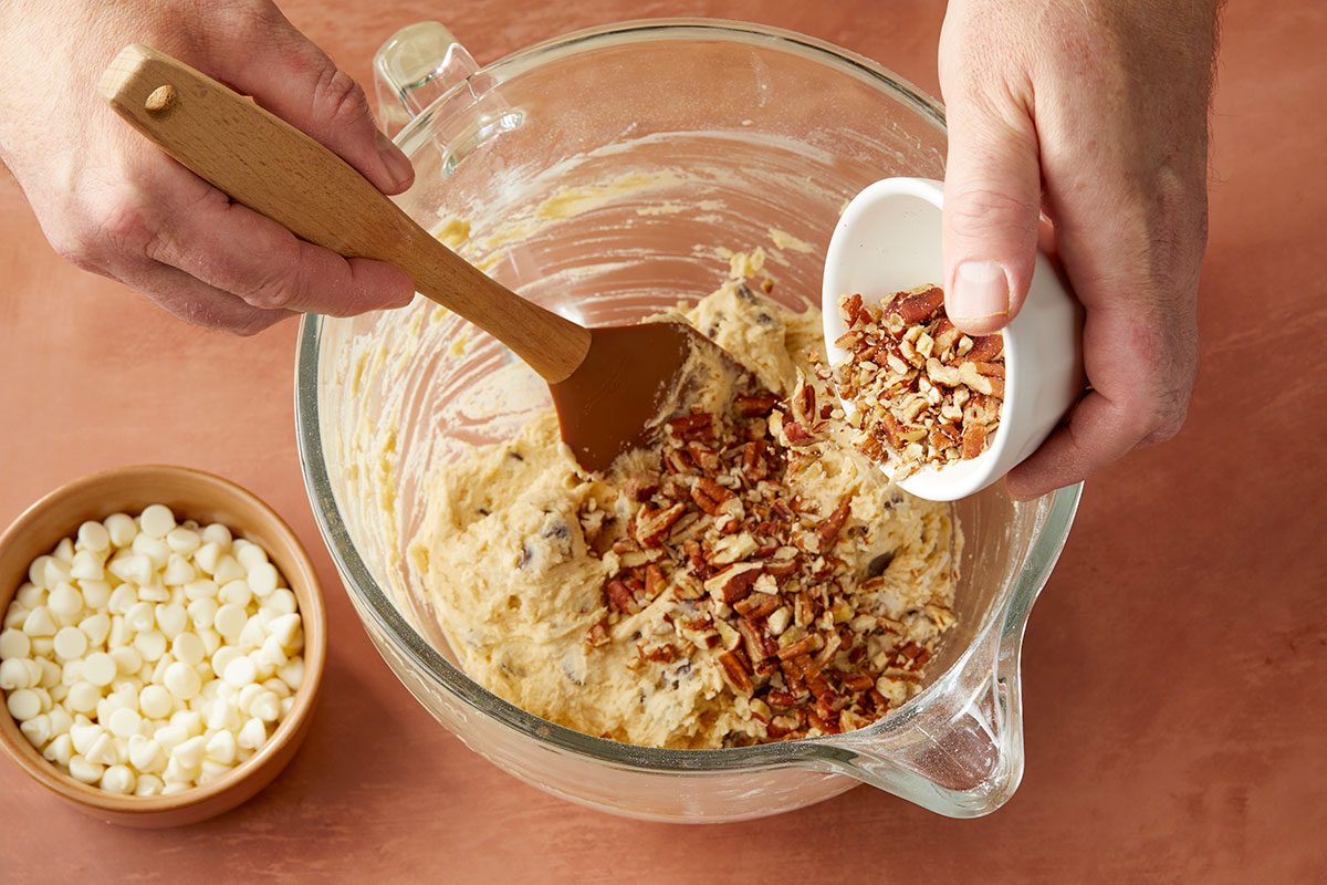 Eye Level horizontal image of chopped pecans being poured from a white ramekin into a clear bowl of batter with a brown spatula resting inside. A bowl of white chips sits next to the mixing bowl. | [Original] Stirring chocolate chips and coconut into batter