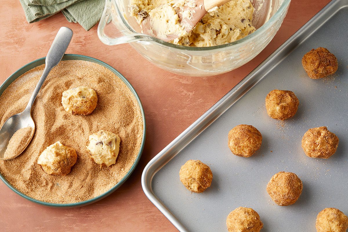 Overhead horizontal image of cookie dough being rolled in cinnamon sugar and placed on a baking sheet. The dough can be seen both in a bowl and after being rolled. Also present is a spoon. | [Original] Process shot - rolling dough into balls, rolling balls in cinnamon sugar, placing balls on baking sheet