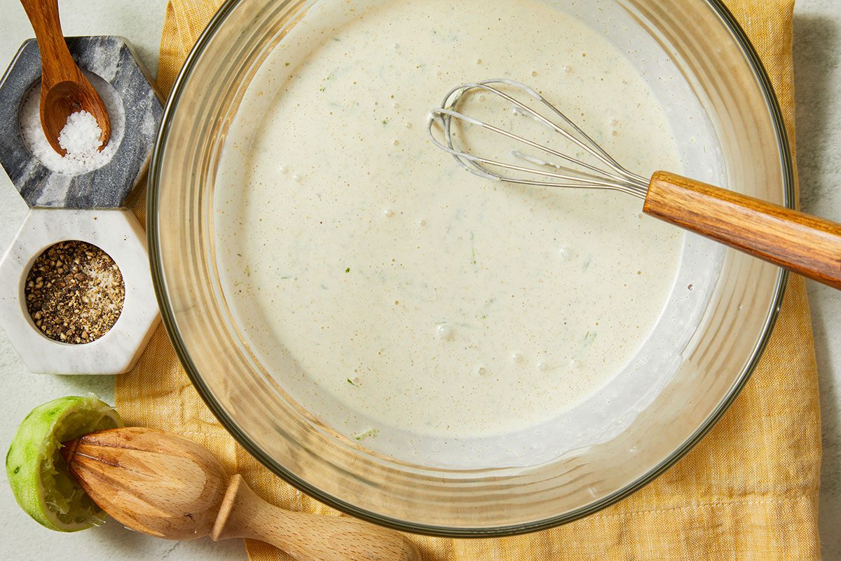 Overhead horizontal image of a clear glass bowl with a white sauce being whisked. The bowl is on a yellow linen towel next to a juiced lime with a citrus reamer and separate bowls of salt and pepper.
