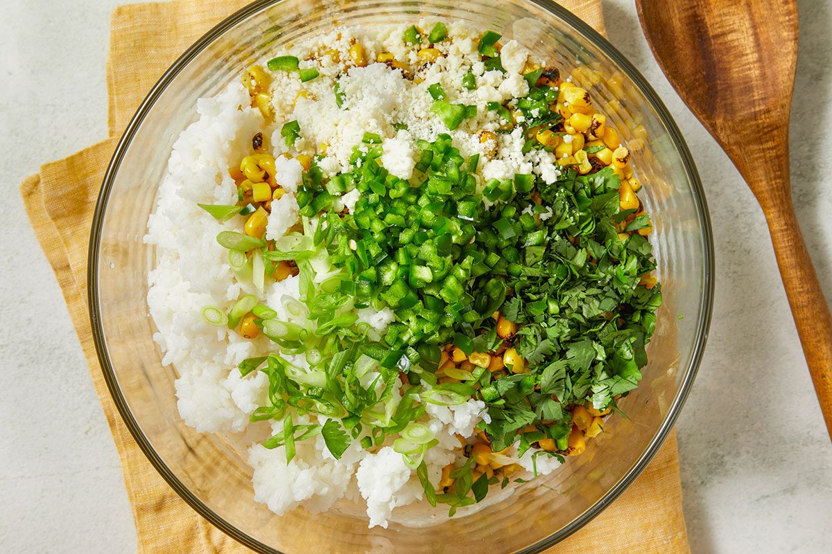 Overhead horizontal image of ingredients in a glass bowl. The bowl contains rice, charred corn, queso fresco, jalapeños, green onion and cilantro.
