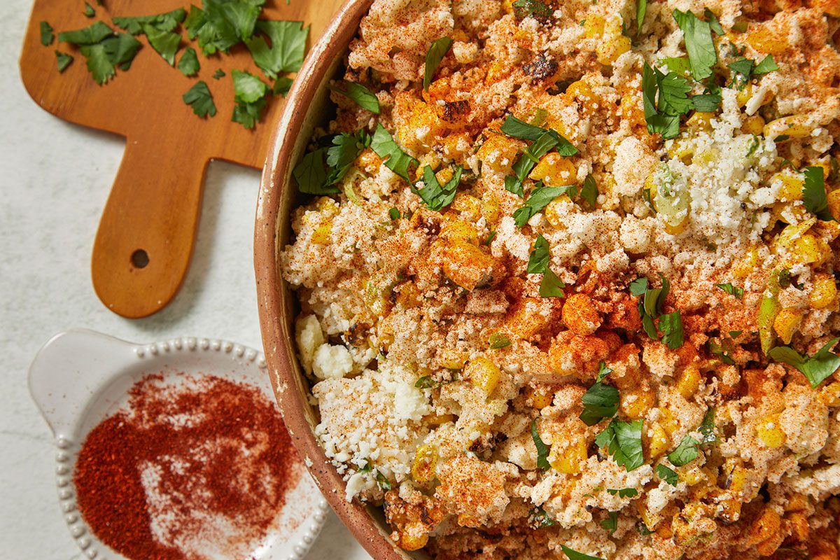 Overhead horizontal image of a dish of Mexican Street Corn Salad, also known as esquites. The dish is topped with crumbled cheese, chili powder, and chopped cilantro, while a small dish of chili powder and a wooden cutting board with chopped cilantro sit next to the bowl.