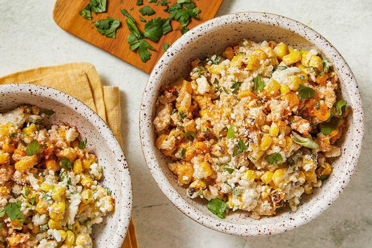 Overhead horizontal image of two bowls of Mexican street corn salad. The salad contains charred corn, cilantro, cheese, and a light dusting of spice.