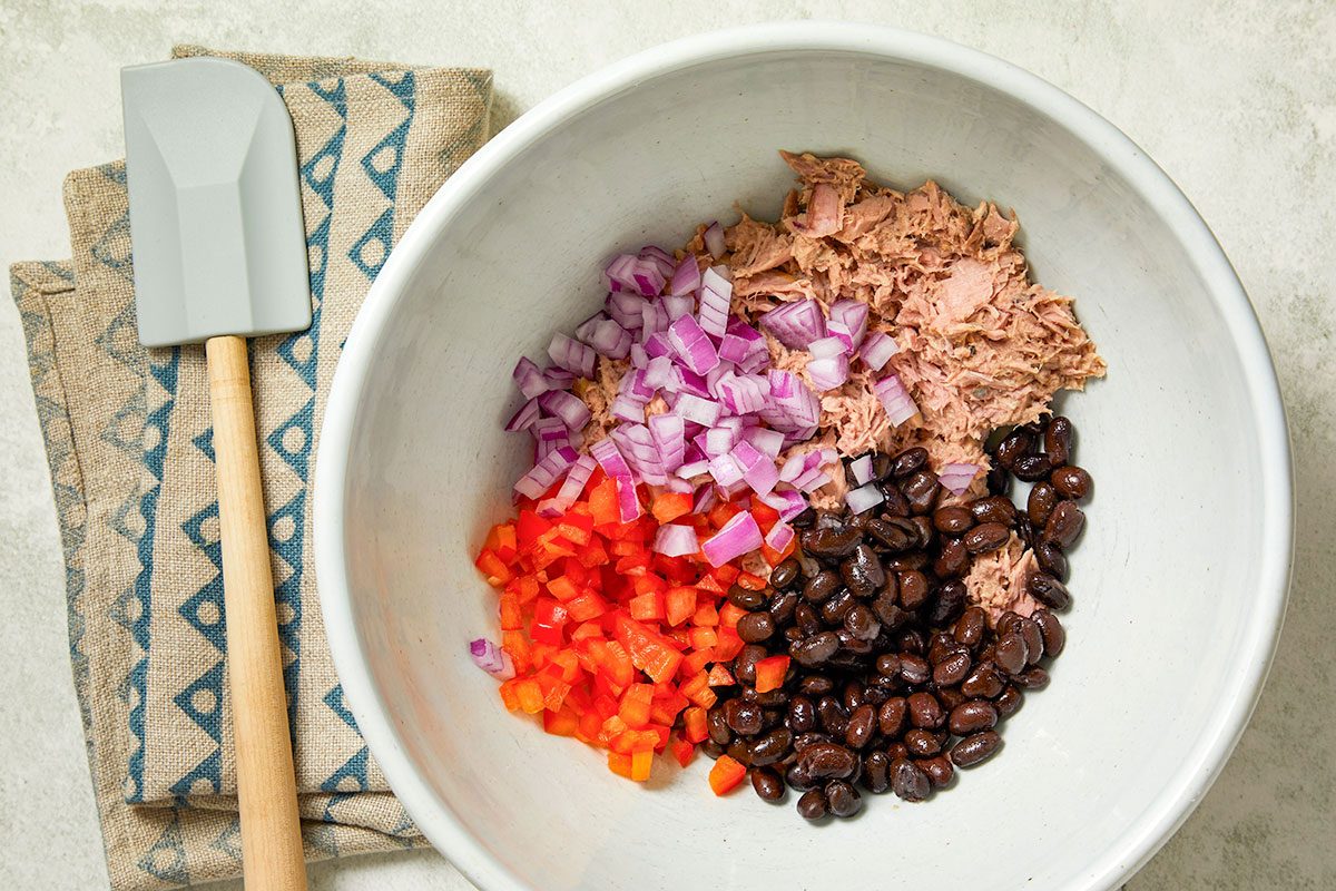 Overhead horizontal image of ingredients in a white bowl. The ingredients, tuna, red onion, black beans and red bell pepper are neatly separated inside the bowl. A gray spatula lays on a patterned linen next to the bowl.
