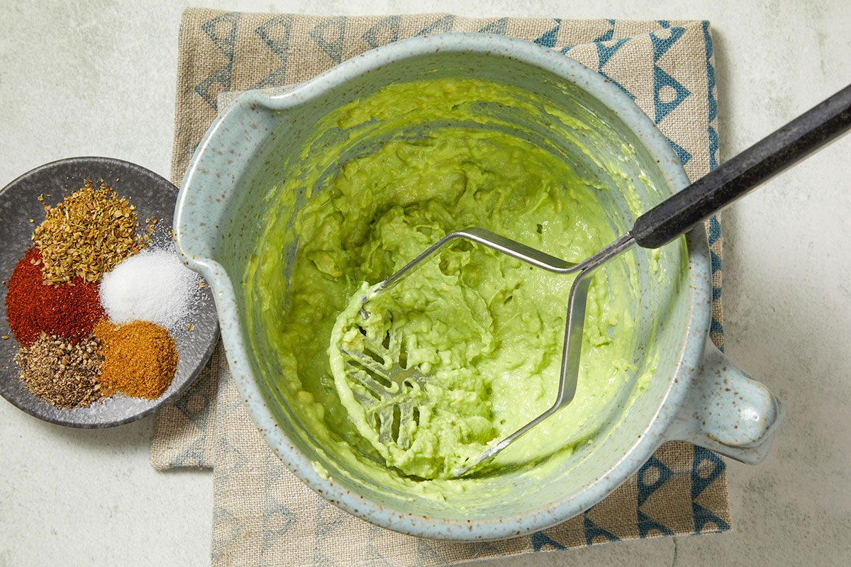 Overhead horizontal image of a light blue bowl filled with mashed avocado. A potato masher is inside the bowl. A small black plate with spices like salt, chili powder, and cumin is to the left of the bowl.