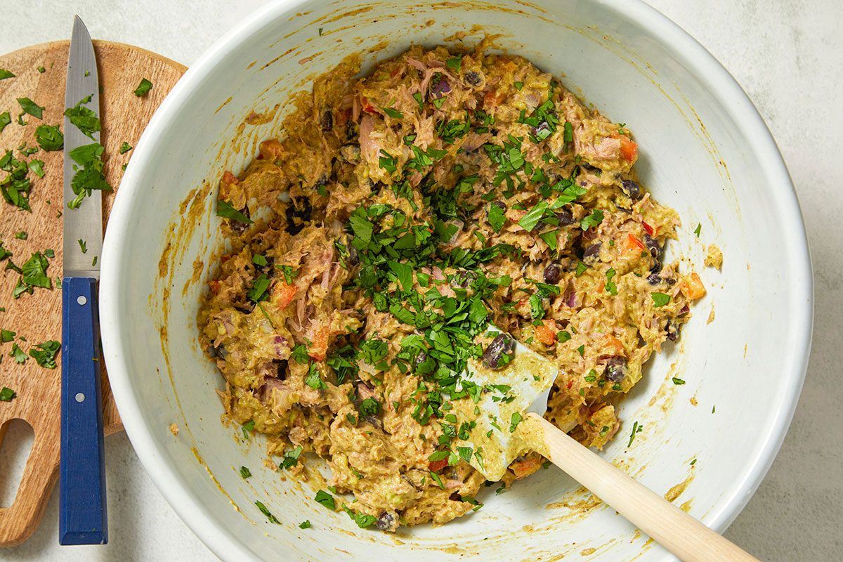 Overhead horizontal image of avocado tuna salad being mixed with a spatula in a white bowl with fresh cilantro on top. Chopped cilantro is visible on a cutting board along with a knife.