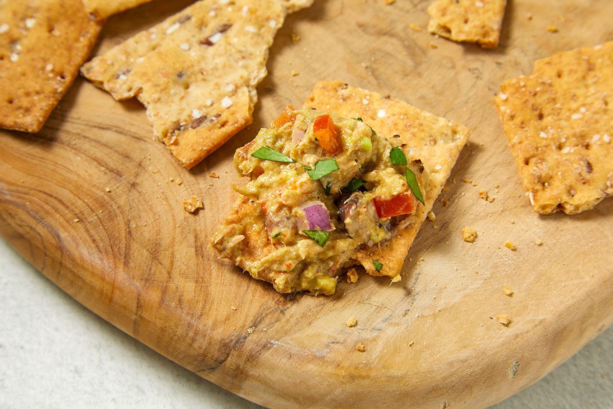 Overhead horizontal image of tuna salad on a cracker, sitting on a wooden board. Red bell pepper and red onion are visible in the tuna salad, which is garnished with fresh basil.