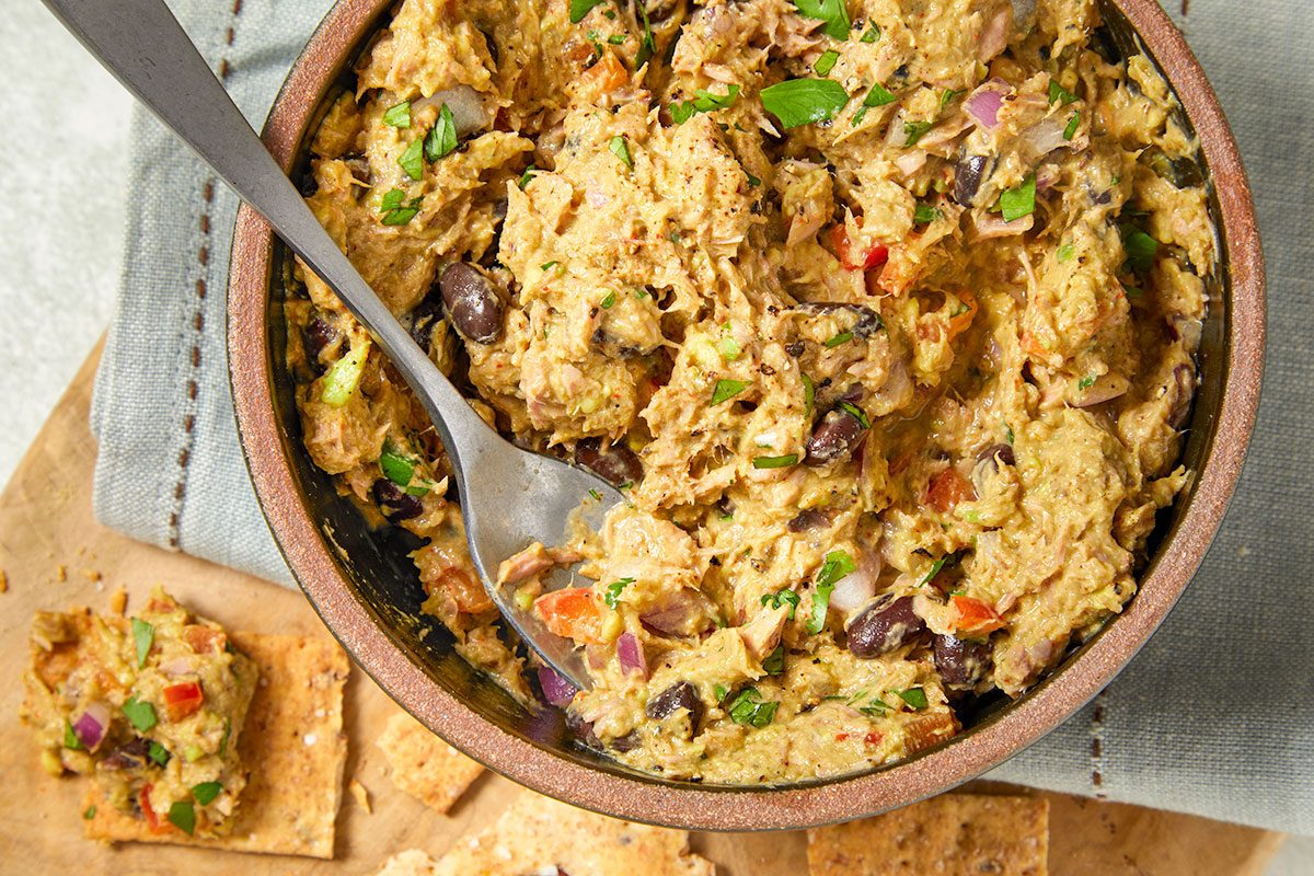 Overhead horizontal image of a bowl of tuna salad with a spoon in the bowl. Visible ingredients include tuna, black beans, red onion, and herbs.