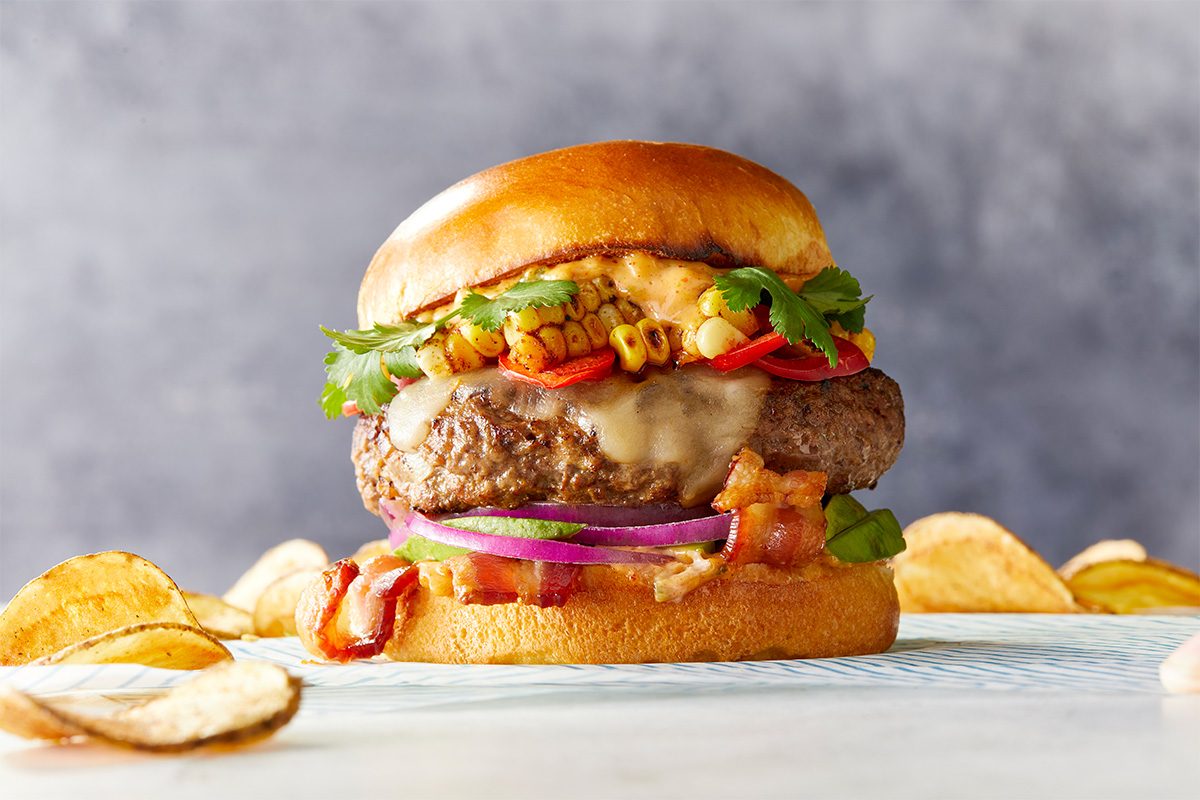 Horizontal image of Festive All-American Hamburgers styled on a light tabletop with potato chips scattered nearby. The burger showcases vibrant layers of grilled corn, bacon, melted cheese, avocado and red onion beneath a glossy toasted bun.