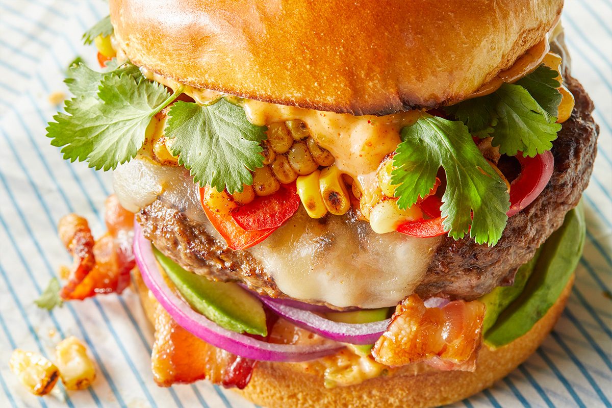 Horizontal close-up detail of a Festive All-American Hamburger highlighting the juicy ground beef patty, melted pepper jack cheese and colorful toppings including grilled corn, bacon, avocado, red onion and fresh cilantro. The tight crop emphasizes texture and freshness.