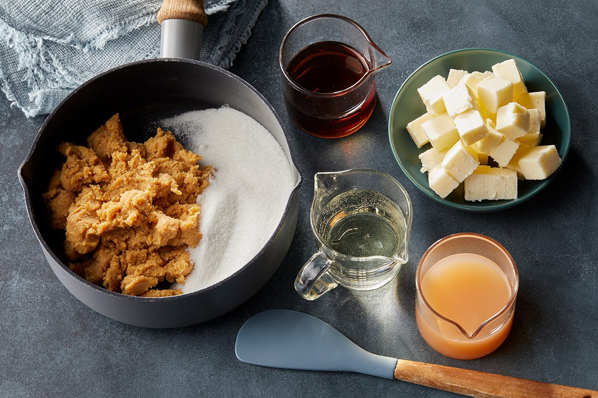 A saucepan with brown sugar and white sugar, surrounded by bowls of cubed butter, light syrup, and apple juice, plus a blue spatula on a dark surface.