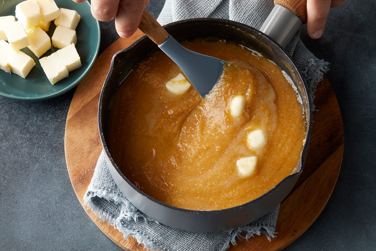 A hand stirs melted butter into a pot of caramel sauce with a spatula, while cubes of butter sit on a plate nearby.