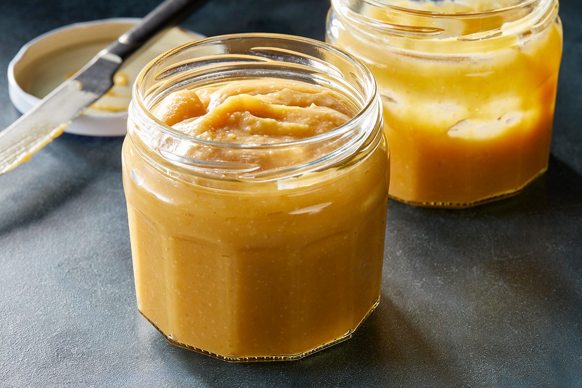 Two glass jars filled with creamy, light brown applesauce sit on a dark surface. A lid and a knife are visible in the background.