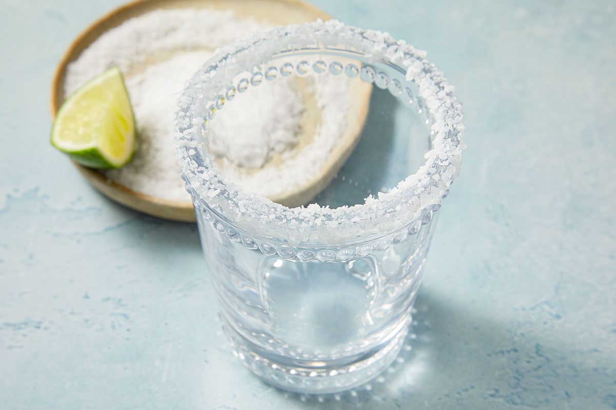 Eye Level horizontal image of a clear glass with a salt rim. Next to the glass is a small plate with salt and a lime wedge.