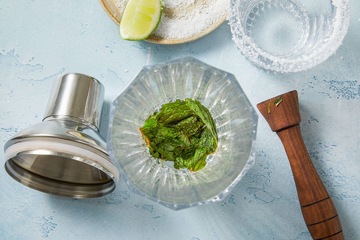 Overhead horizontal image of mint leaves inside of a clear glass, a cocktail shaker, muddler, lime, salt on a small wooden plate, and a glass with a salted rim. The ingredients are arranged on a light blue textured surface.