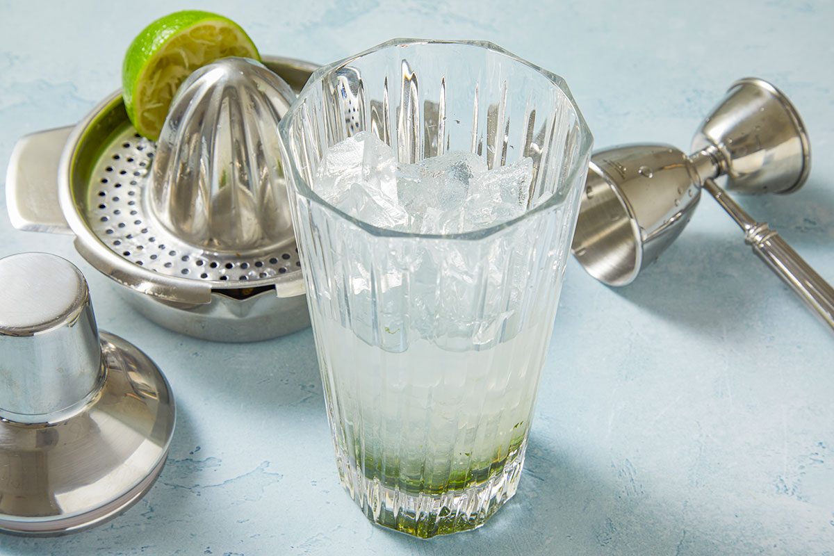 Eye Level horizontal image of the cocktail making process. The image shows a cocktail glass filled with ice, a lime in a citrus squeezer, a jigger, and a cocktail shaker.